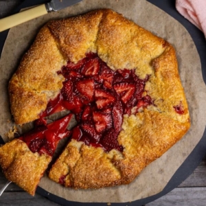 A vertical photo of a sourdough discard strawberry galette brimming with red strawberries. A slice has been taken out of the galette, but is still sitting on the plate. There is a bowl of vanilla ice cream sitting on a pink linen napkin above the galette and a small jar of strawberry sauce.