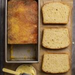 Sourdough English muffin loaf on parchment paper with three slices of bread next to a bowl of butter with a butter knife.