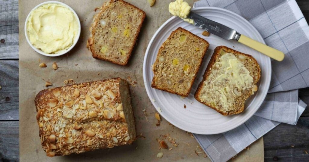 Sourdough Hawaiian banana bread on parchment paper with half the loaf sliced and two slices on a white plate with butter.