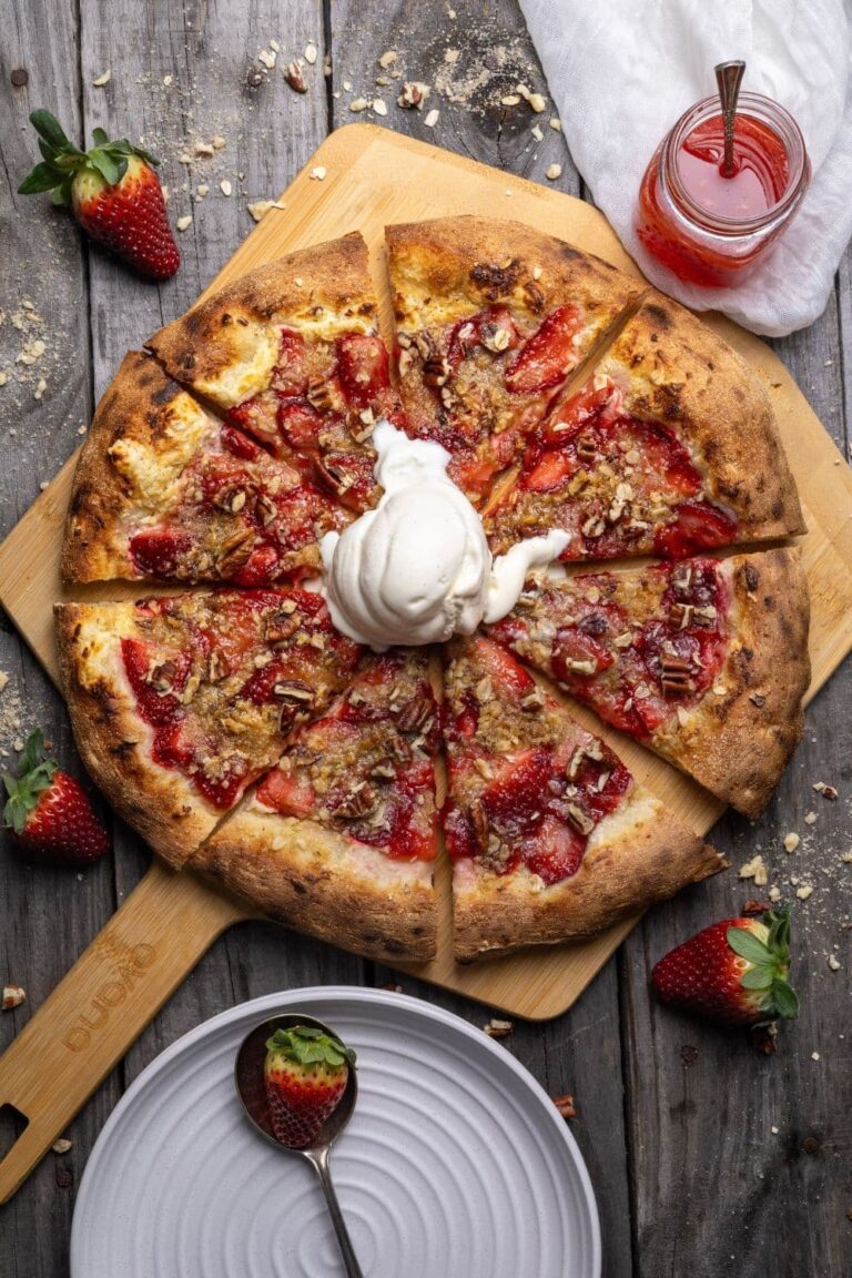 A sourdough strawberry crumble pizza topped with vanilla ice cream and sliced into 8 slices. There is a small jar of strawberry sauce and some fresh strawberries in the photo too.