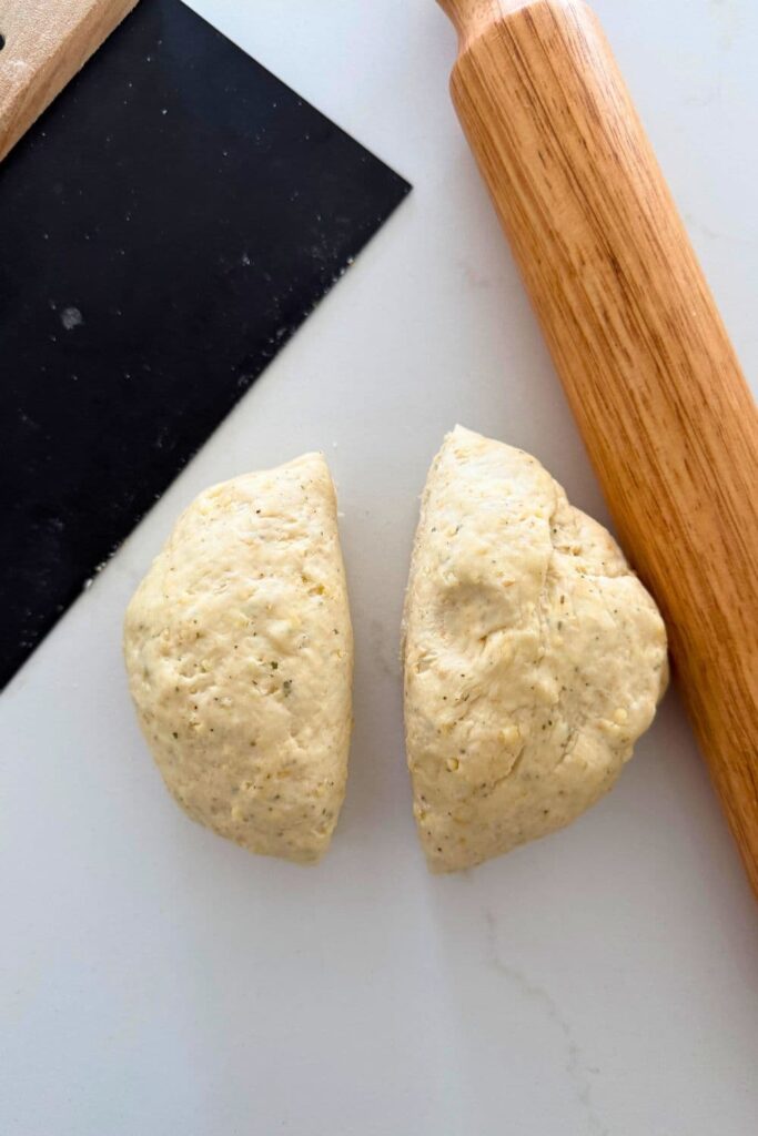 Two portions of sourdough cracker dough sitting on the counter top with a black dough scraper and rolling pin.