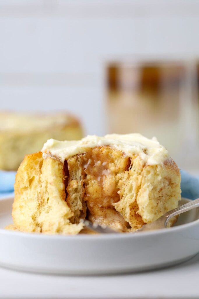 A sourdough cinnamon roll sitting on a white plate. The cinnamon roll is filled with gooey cinnamon brown sugar filling and topped with vanilla cream cheese filling. You can see a cup of coffee in the background, as well as a light blue napkin.
