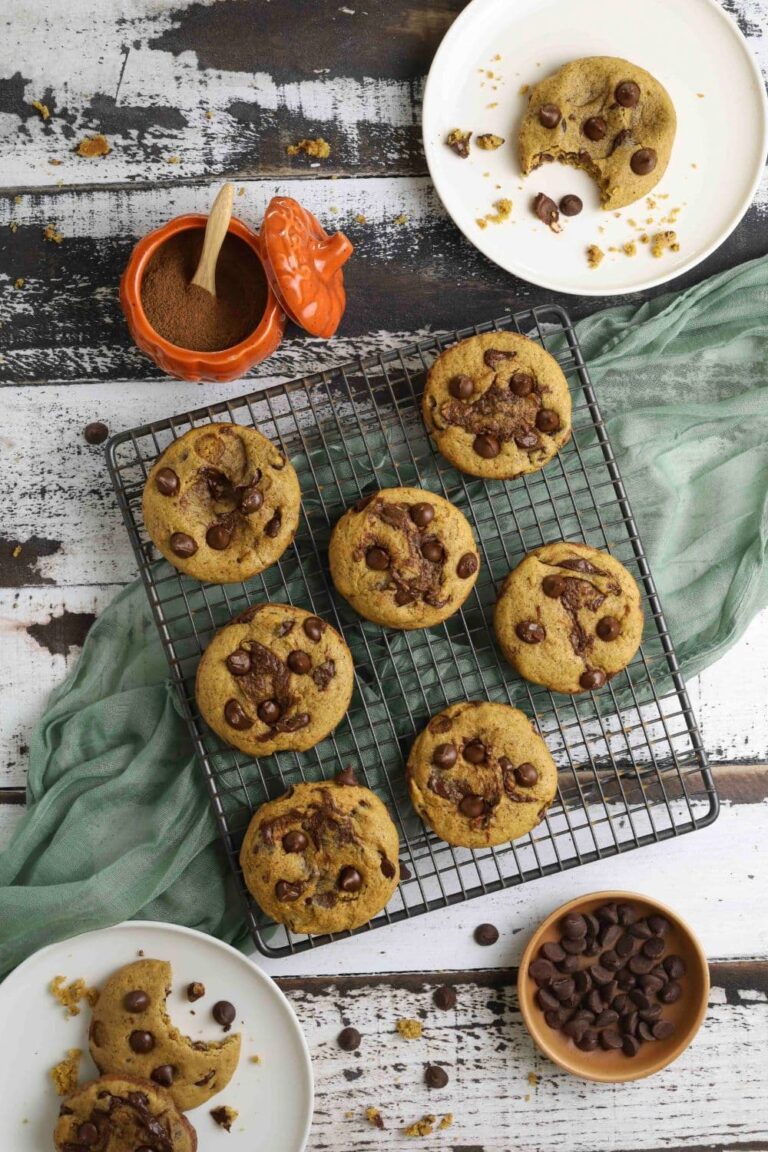 Sourdough pumpkin chocolate chip cookies on a cooling rack next to pumpkin spices and plates of partially eaten cookies.