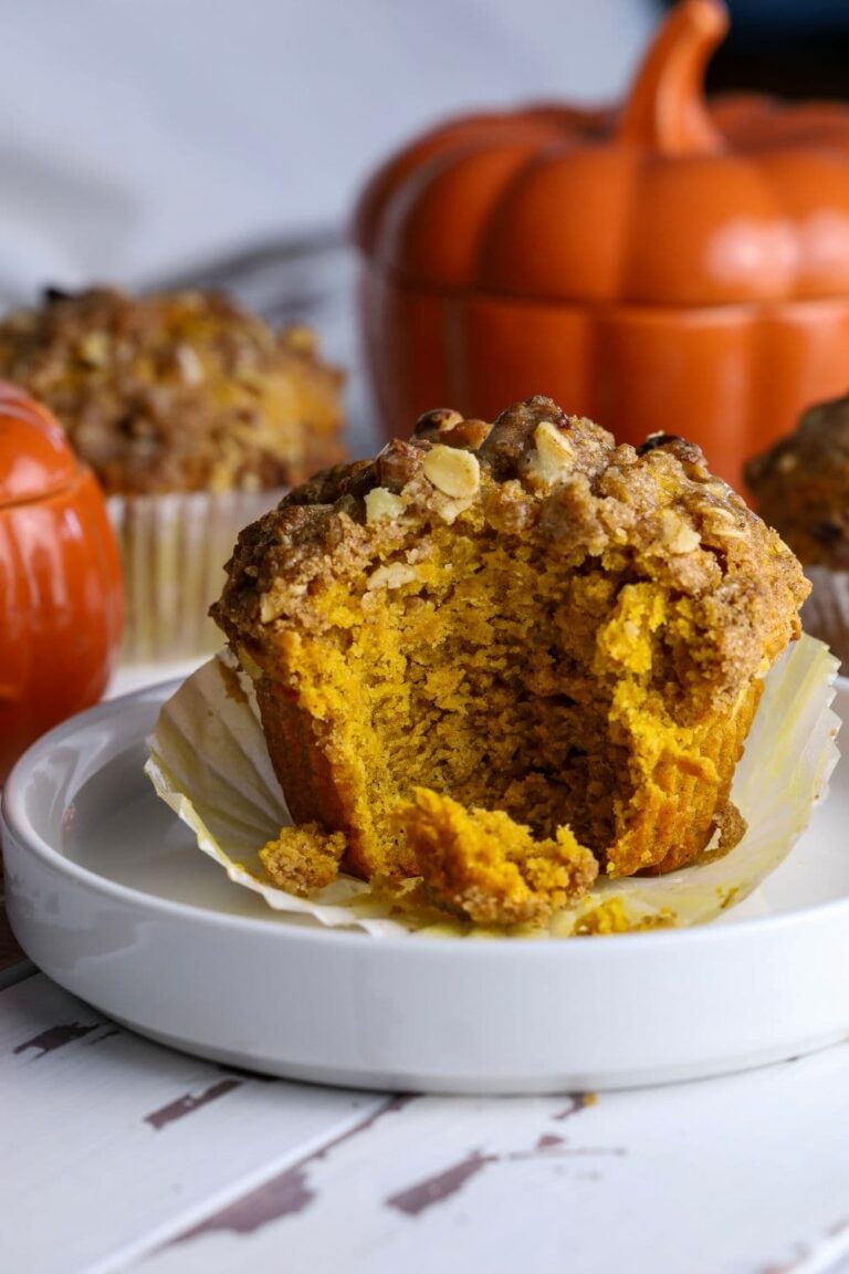A photo of a sourdough pumpkin muffin that has a bite taken out of it so you can see the tender crumb and rich pumpkin color inside. There is an orange pumpkin in the background.
