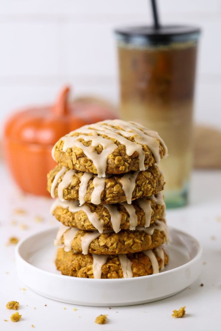 A stack of 5 sourdough pumpkin oatmeal cookies topped with maple glaze. They are on a white plate in front of a ceramic pumpkin and iced coffee.