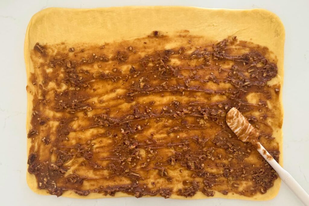 Sourdough maple pecan roll dough on counter with a spatula spreading filling on top.