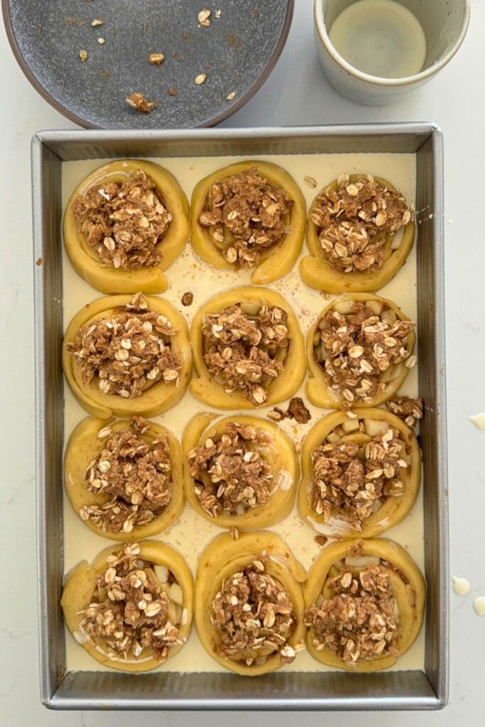 Sourdough apple cinnamon roll dough slices in baking dish with heavy cream poured in and streusel topping on top of dough.