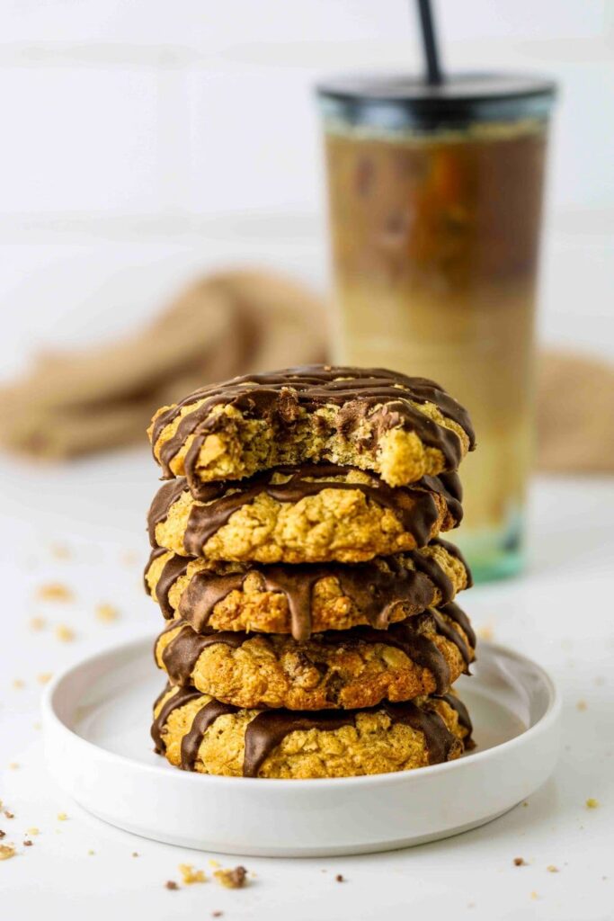 Sourdough Oatmeal Chocolate Chip Cookies stacked on a plate with a bite taken out of top cookie and an iced coffee in the background to drink with the cookies.