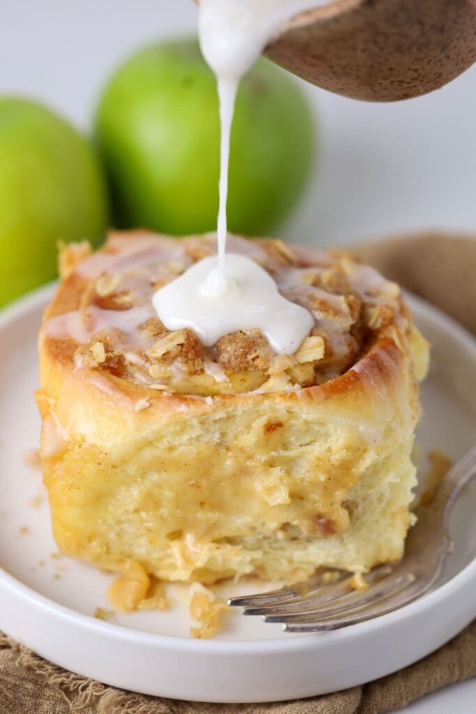 A sourdough apple cinnamon roll topped with streusel and drizzled with vanilla glaze. There are two apples in the background.