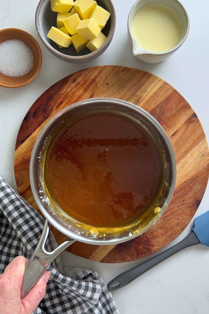 A saucepan filled with granulated sugar that has been being melted into an amber liquid. You can see a jug of cream, butter and dish of salt in the photo as well.