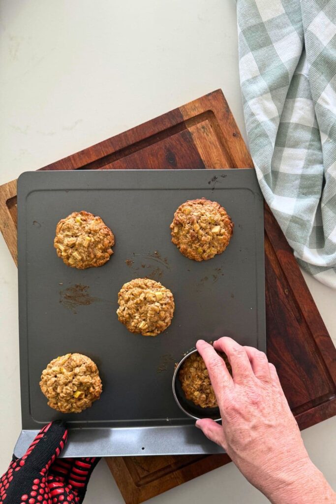 Fresh baked apple cinnamon sourdough oatmeal cookies on a cookie sheet with a hand to correct cookie edges, making them round.