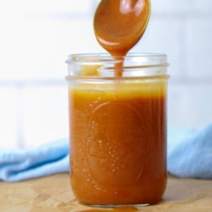A glass mason jar filled with homemade caramel sauce being drizzled back into the jar with a spoon.