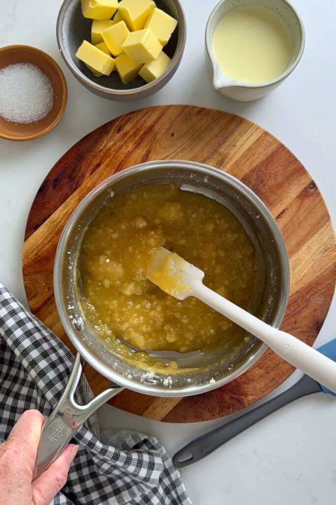 A saucepan filled with granulated sugar that is being melted. The sugar has started to melt quite a bit and has become semi liquefied. You can see a jug of cream, butter and dish of salt in the photo as well.