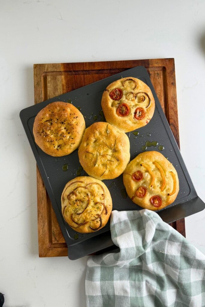Fresh baked sourdough focaccia rolls on a baking sheet sitting on top of a wooden cutting board to cool.