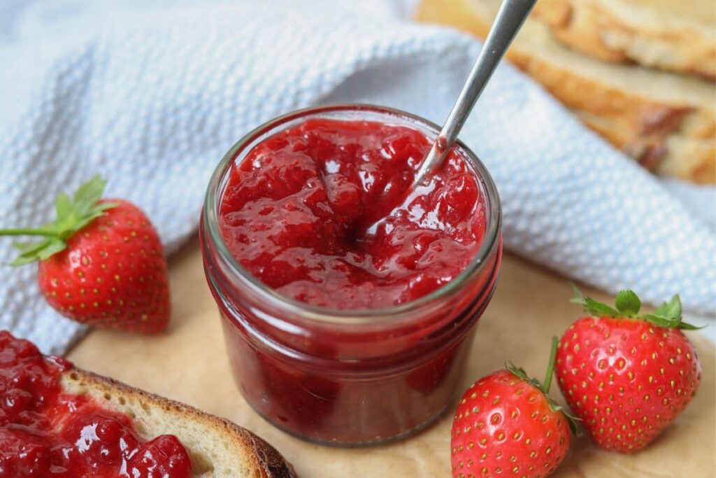 A small jar of strawberry jam with a spoon in it surrounded by fresh strawberries and some slices of sourdough bread.
