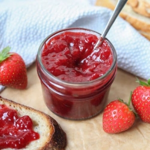 A small jar of strawberry jam with a spoon in it. There are fresh strawberries scattered around it.