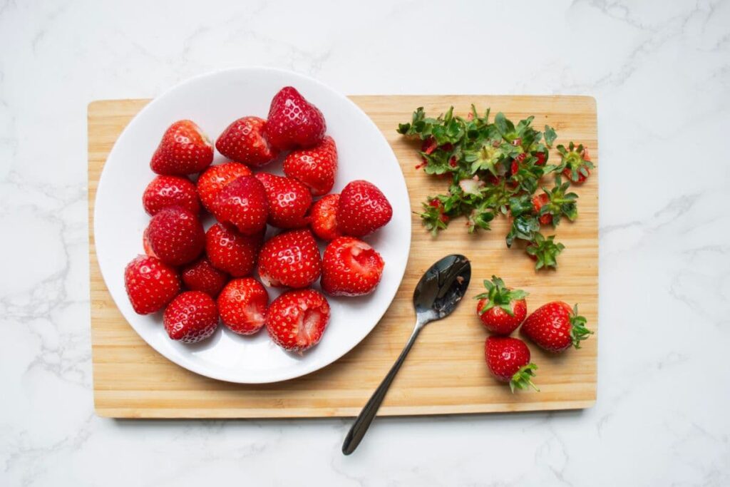 A wooden chopping board holding a whit plate with strawberries that have been hulled using a spoon.