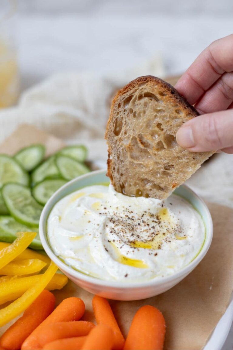 A bowl of whipped feta dip topped with olive oil and surrounded by colorful veggies. A piece of whole wheat sourdough bread is being dipped into the bowl of dip.