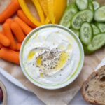A bowl of whipped feta dip topped with olive oil and black pepper. The bowl has been served with carrots, yellow peppers, cucumber slices, black olives and whole wheat sourdough bread slices.