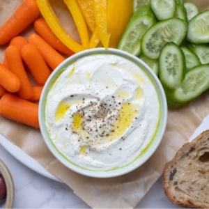 A bowl of whipped feta dip topped with olive oil and black pepper. The bowl has been served with carrots, yellow peppers, cucumber slices, black olives and whole wheat sourdough bread slices.