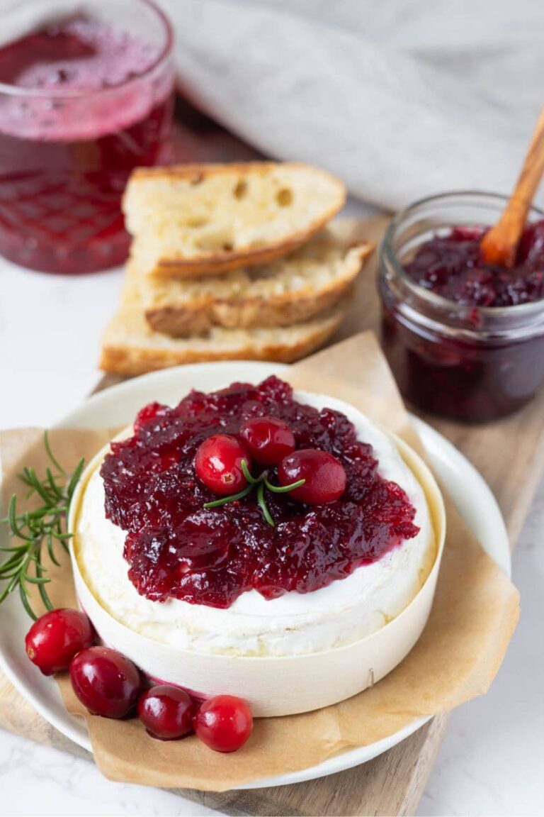 A wheel of Brie topped with vibrant cranberry vanilla jam. The Brie is decorated with fresh cranberries and rosemary and you can see the jar of jam and stack of sourdough bread slices in the background.