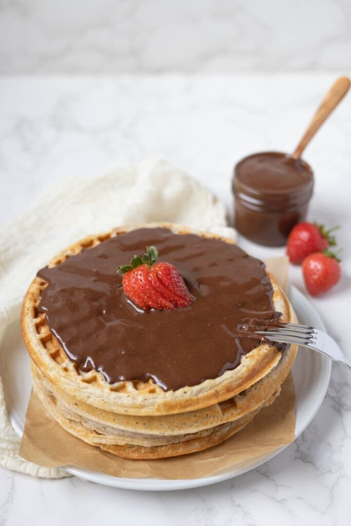 Waffles stacked on a plate with homemade Nutella and strawberries on top.
