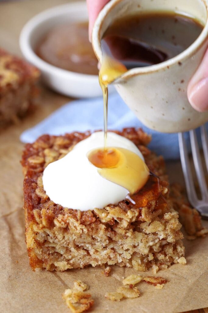 Pouring maple syrup over a slice of sourdough baked oatmeal that's topped with yogurt.