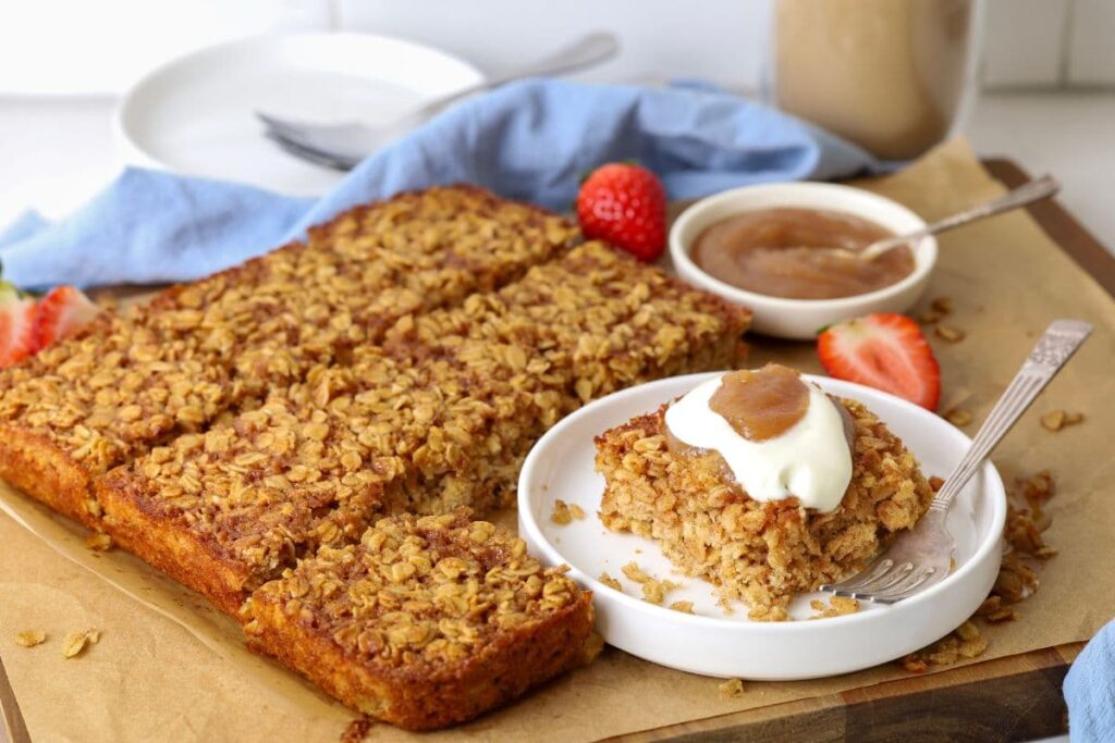 Sliced sourdough baked oatmeal in squares on parchment paper with one slice on a plate topped with yogurt and apple butter for serving.
