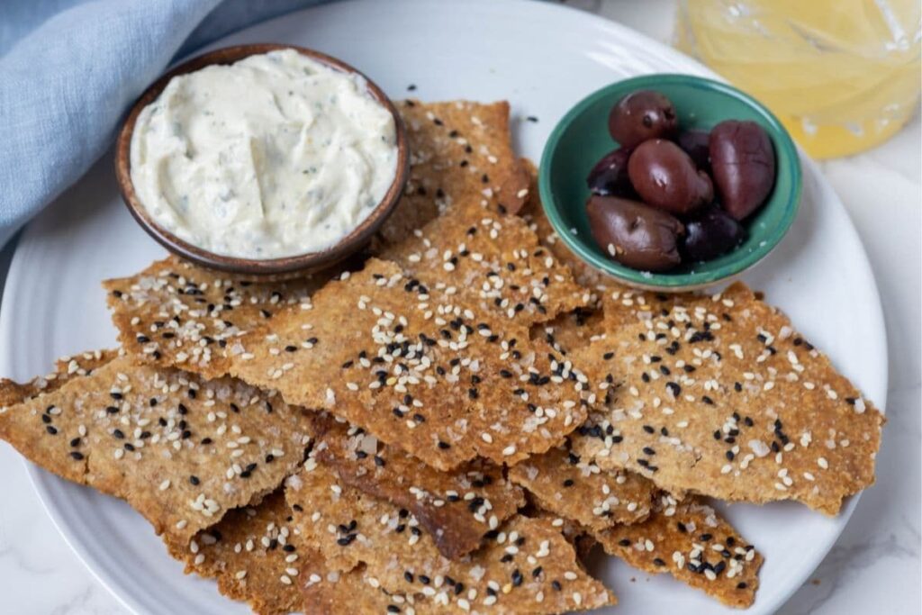 A horizontal image of some sourdough lavosh crackers displayed on a plate with some dip and black olives in a small green bowl.