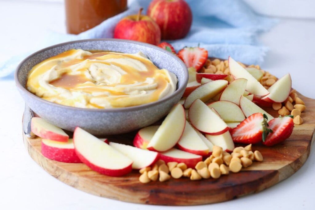 A bowl of whipped caramel dip on a wooden tray next to sliced apples and strawberries.