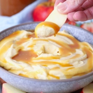 Close up of a hand dipping an apple slice into a bowl of whipped caramel dip.